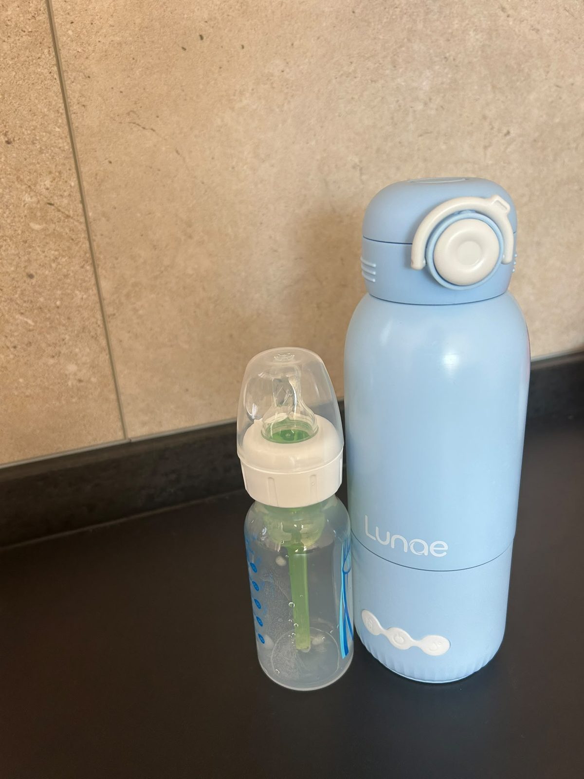 Lichtblauwe Lunae draagbare flessenwarmer naast een babyfles op een zwart aanrecht