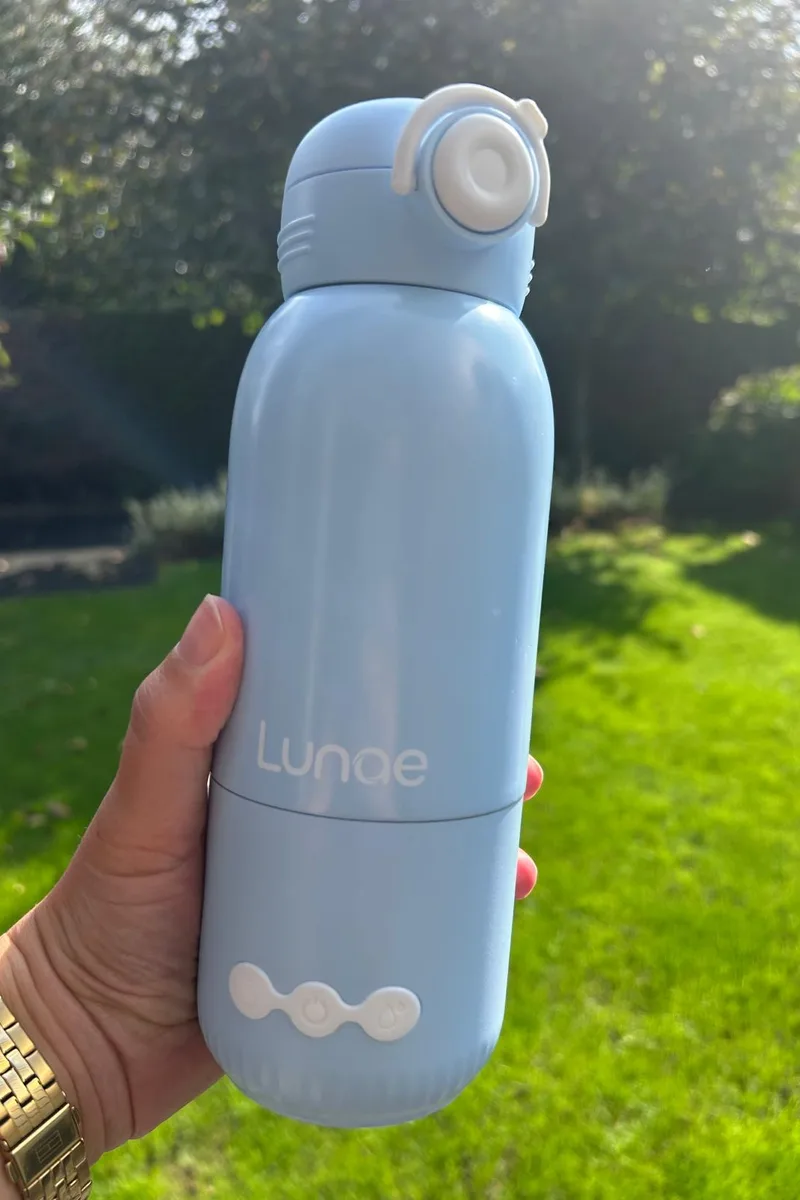 Hand houdt een lichtblauwe Lunae draagbare flessenwarmer vast in een zonnige tuin