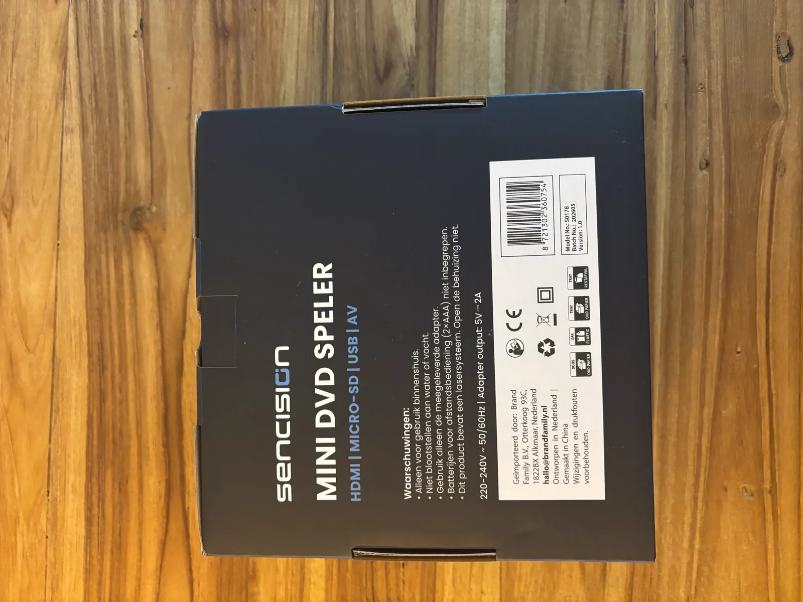 Achterkant van de doos van de Sencision mini dvd-speler met waarschuwingen, adaptergegevens en de importeur Brand Family uit Alkmaar