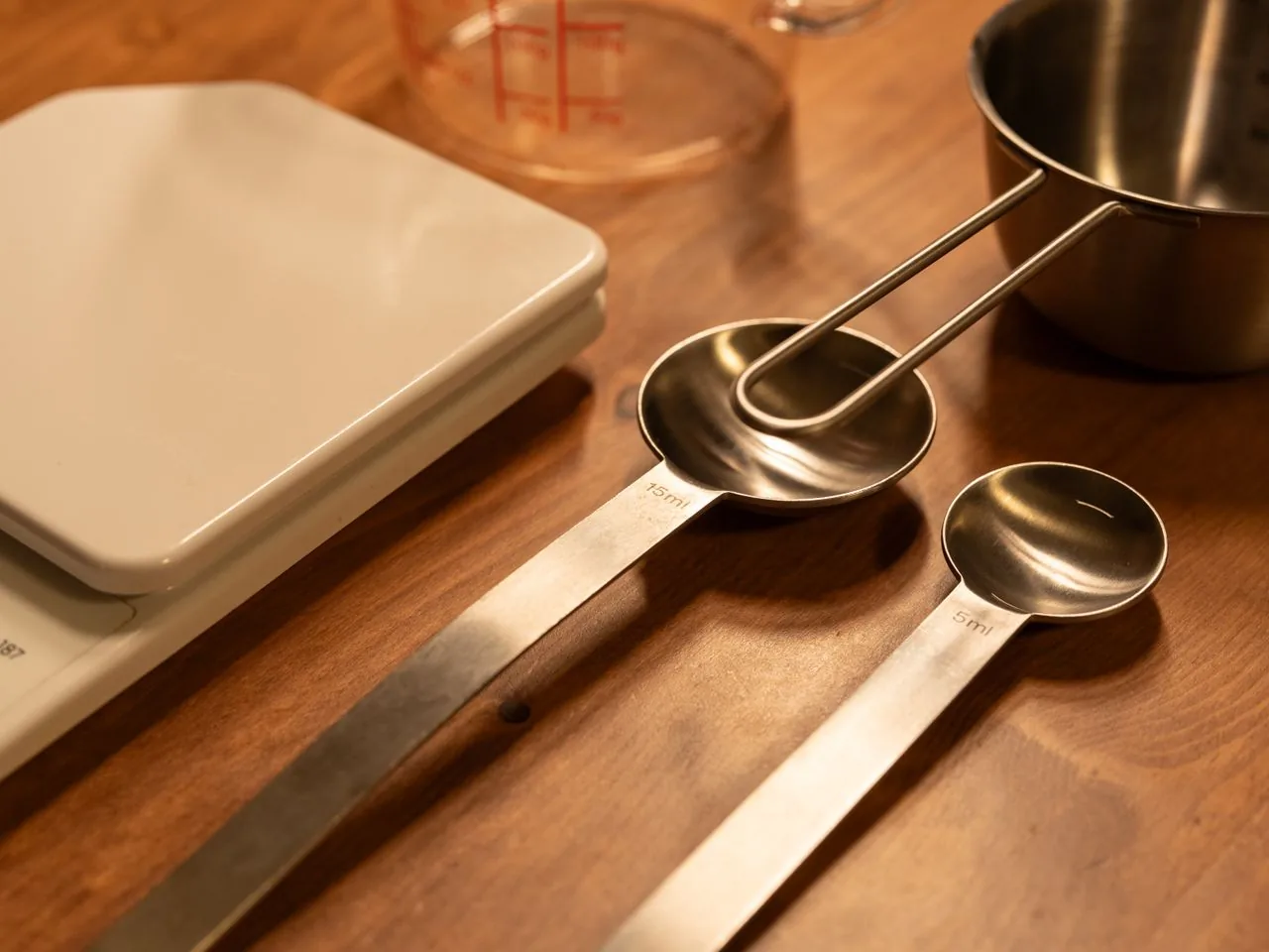 Witte digitale keukenweegschaal met metalen maatlepels en een RVS maatbeker op een houten tafel