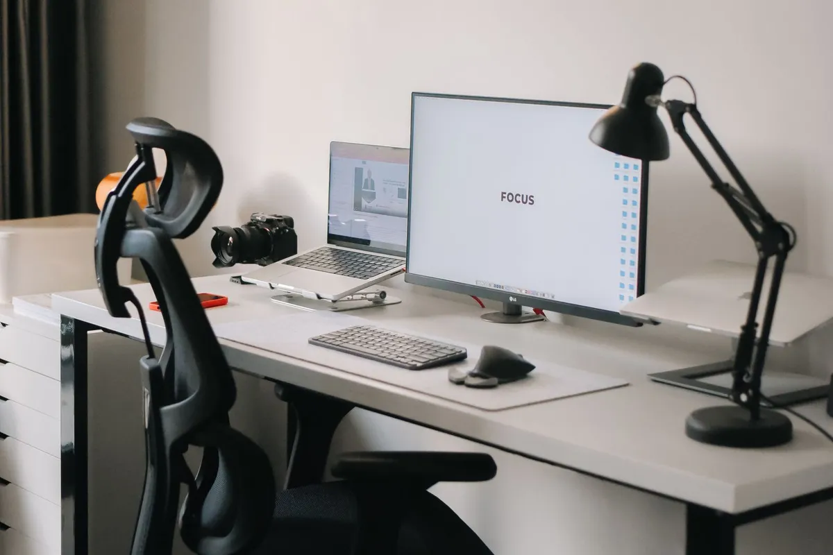 Ergonomische bureaustoel aan een wit bureau met monitor, laptop en bureaulamp in een lichte thuiswerkplek